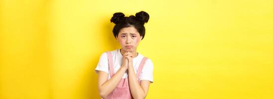 Sad asian girl pleading for help, begging you, sulking and whining, standing on yellow background photo