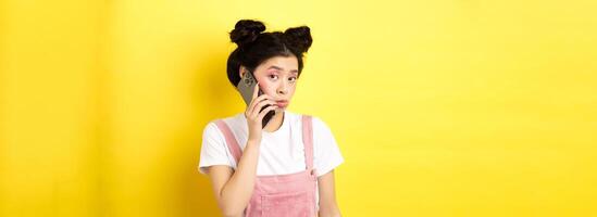 Cute teen girl talking on smartphone, making silly pouting face and look timid at camera, standing with glamour makeup on yellow background photo