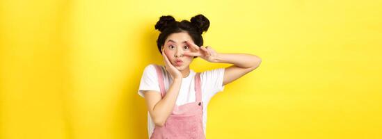 Kawaii asian girl showing v-sign and pouting cute, making silly face with makeup, standing on yellow background photo