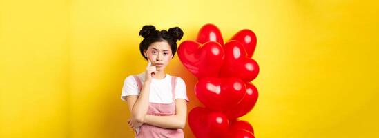 Valentines day concept. Unamused and bored asian girlfriend staring skeptical at camera, waiting for something near red hearts balloons, yellow background photo