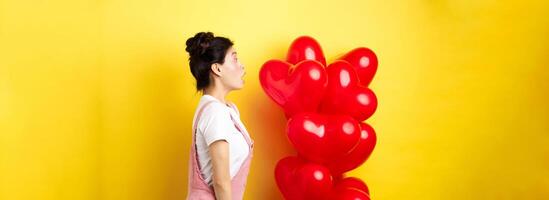 Valentines day and relationship concept. Profile of surprised asian girl staring left at logo, standing near heart balloons, yellow background photo
