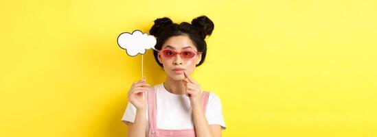 Summer and fashion concept. Stylish asian girl in sunglasses holding comment cloud on stick and looking thoughtful, thinking or saying something, standing on yellow background photo
