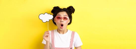 Summer and fashion concept. Excited party girl in sunglasses, holding comment cloud party mask and gasping amazed, standing impressed on yellow background photo