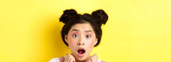 Head shot of shocked asian girl with hairbuns and glamour makeup, open mouth and looking startled at camera, standing against yellow background photo