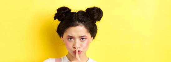 Close up portrait of angry asian girl shushing, press fingers on lips and tell be quiet, scolding loud person, need silence, telling big secret, standing on yellow background photo