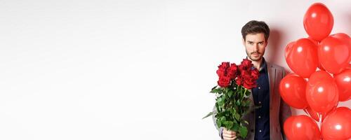Macho man going on romantic date, looking confident at camera, holding gifts for lover on Valentines day, standing with heart balloons and red roses, white background photo