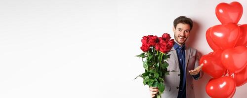 Romantic guy in suit saying happy Valentines day, giving red roses to you and pointing hand, standing with heart balloon gift over white background photo