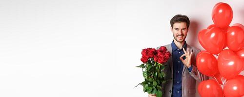 Young man in suit have all under control, showing OK sign and holding red roses, prepare romantic surprise with heart balloons and flowers on Valentines day, white background photo