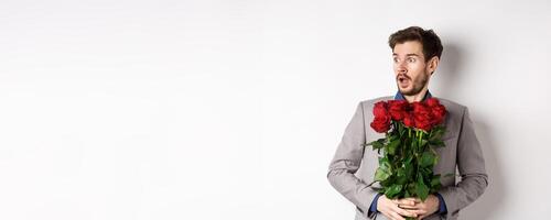 Handsome young man in suit holding red roses, looking left with surprised and startled expression, standing on Valentines day over white background photo