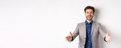 Happy handsome man in suit stretch out hands to greet business partners, welcome guests and smiling excited, standing on white background photo