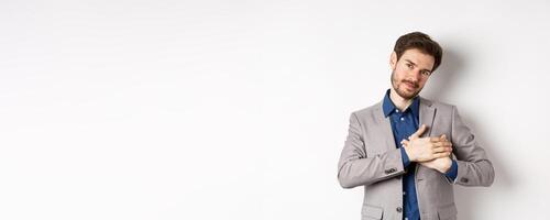 Romantic handsome guy with beard holding hands on heart and smiling, thanking you, feeling nostalgic, having tender memories in heart, white background photo