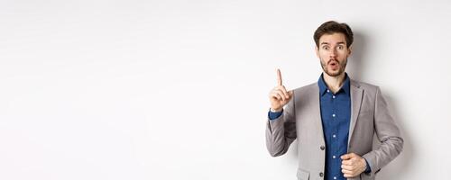 Excited businessman in suit pitching good idea, raising finger in eureka sign and looking amused, saying suggestion, standing on white background photo