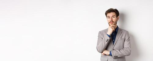 Thoughtful businessman in suit touching beard, thinking and looking at camera, deciding with pensive face, standing on white background photo