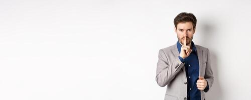 Angry businessman showing taboo sign, shushing and tell to keep quiet, need silence, standing with finger on lips and looking at camera, white background photo