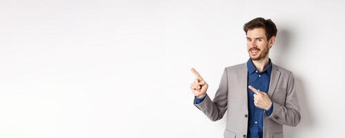 Handsome bearded businessman in grey suit pointing fingers left at logo, inviting check out advertisement, standing on white background photo
