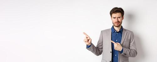Bad idea. Displeased skeptical guy in business suit smirk and frown, pointing fingers left at upsetting news, looking hesitant at camera, white background photo