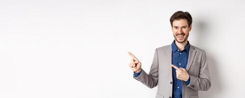 Handsome caucasian man in business suit pointing fingers left at banner and smiling, showing advertisement, standing against white background photo