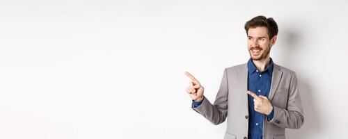 Successful businessman in grey suit pointing fingers left and looking at banner, smiling confident, showing advertisement, standing against white background photo