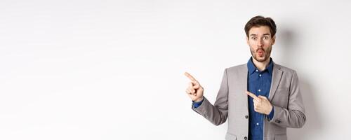 Excited handsome man in suit pointing fingers left at banner and saying wow, look here gesture, showing advertisement, standing on white background photo