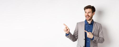 Excited bearded guy in festive suit pointing and looking left, smiling amazed, showing cool promo deal, standing on white background photo