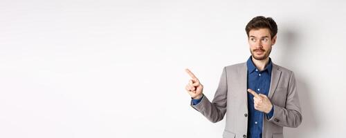 Doubtful male entrepreneur in suit looking and pointing left with hesitant face, standing unsure against white background photo