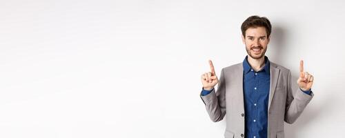 Confident successful businessman pointing fingers up, smiling and showing company logo, standing in suit on white background photo