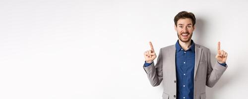 Excited lucky businessman in suit pointing fingers up, smiling amused, showing cool advertisement, standing on white background photo