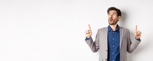 Amazed bearded guy in business suit pointing and looking up wondered, say wow and showing exciting thing, standing against white background photo