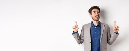 Excited handsome man with beard, wearing suit and looking amused up, pointing at top with admiration, standing against white background photo