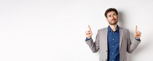 Skeptical and hesitant young businessman in suit looking up with disappointed smirk, feel unsure about deal, standing dissatisfied on white background photo