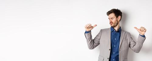 Handsome and confident man in suit pointing at himself, feeling cool and sassy, self-promoting his good appearance, standing on white background photo