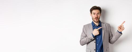Excited handsome caucasian businessman in suit pointing right at logo, say wow and look amazed, standing on white background photo