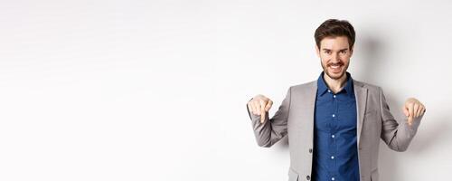 Smiling handsome man in business suit pointing fingers down, showing good deal, look here gesture, standing on white background photo