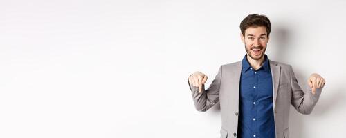 Hey check this out. Happy businessman in suit pointing fingers down and smiling, inviting to event, showing link, standing on white background photo