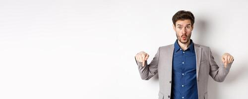 Wow look down. Excited business guy in suit pointing fingers bottom advertisement and look amazed, standing on white background photo