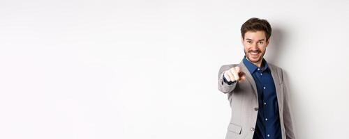 Its you. Smiling handsome manager in suit pointing at camera, choosing person and looking pleased, standing on white background photo