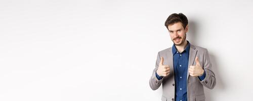 Very good. Smiling handsome male ceo manager showing thumbs up in approval, look proud and satisfied, say yes, approve nice work, standing on white background photo