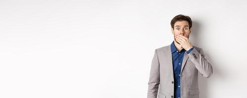 Shocked boyfriend in suit gasping, covering mouth with hand and pop eyes at camera startled, standing on white background photo