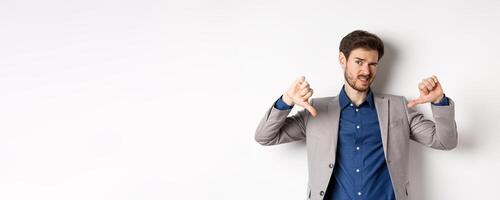 It sucks. Man feeling awkward showing thumbs down and judging something bad, frowning disappointed, standing in suit on white background photo