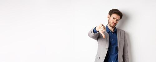 Negative emotions. Disappointed businessman frowning, showing thumb down to disapprove product, dislike something bad, stading on white background photo