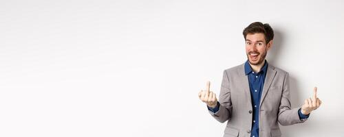 Rude ignorant guy in business suit showing middle fingers and tongue, smiling while mocking people, fuck you gesture, standing on white background photo