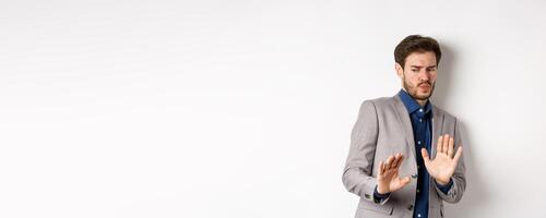 Stay away. Reluctant businessman step back with concerned disgusted face, raising hands to block bad offer, rejecting something awful, tilting from disgust, white background photo