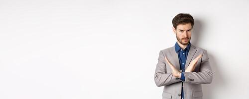 Serious business man making cross and says no, looking determined, disagree and prohibit something bad, tell to stop, standing in suit on white background photo
