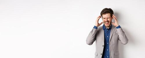 Frustrated business man in suit going crazy, screaming and shaking hands near head, feeling annoyed or angry, shouting distressed, standing on white background photo