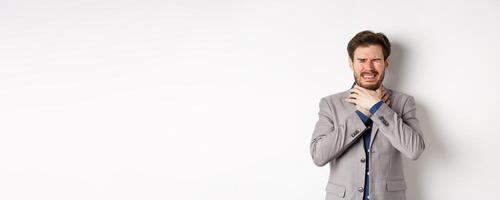 Crying man in suit holding hands on neck and look distressed, feel sick with sore throat, standing on white background photo
