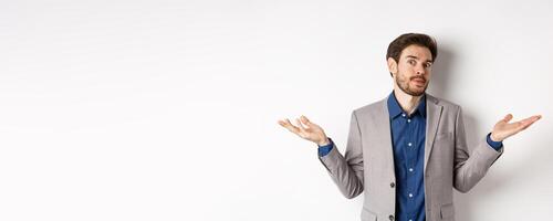 I dont know. Clueless bussiness man in suit shrugging shoulders and looking unaware, have no idea, standing on white background photo
