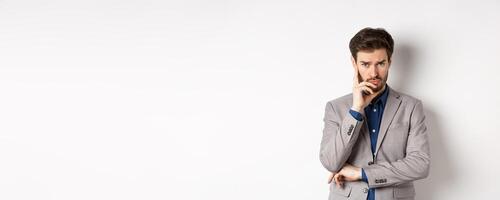 Troubled frowning business man in suit looking at camera pensive, thinking or making decision, standing on white background photo