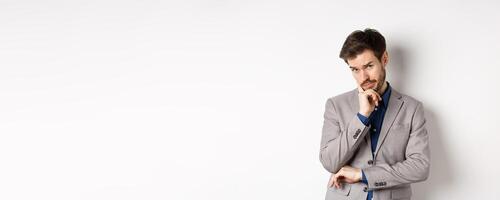 Hesitant businessman in suit making decision, looking pensive at camera with concerned face, having doubts, standing on white background photo