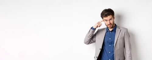 Annoyed male employer scolding personal, pointing finger at head, are you stupid out of your mind gesture, stare at camera from under forehead, standing in suit on white background photo