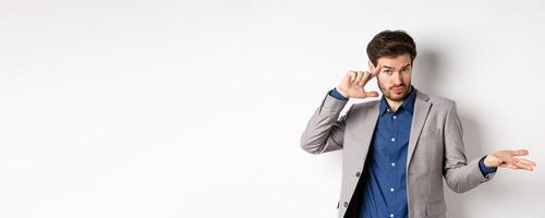 Are you crazy. Young man in suit pointing finger at head to scold strange or stupid mistake, shrugging shoulders confused, standing on white background photo